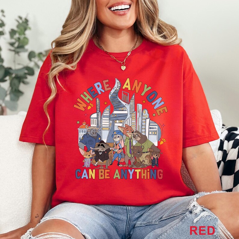 May include: Red t-shirt with a colorful graphic featuring cartoon characters and the text "WHERE ANYONE CAN BE ANYTHING." The design includes a cityscape and various animal figures. The shirt is a casual, oversized fit.