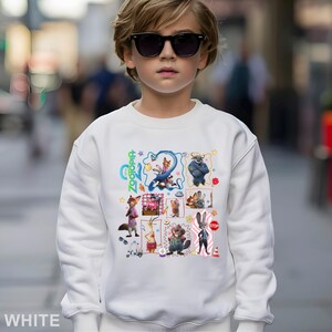 May include: White sweatshirt featuring a colorful graphic of characters from the animated film "Zootopia." The design includes images of a fox, a bunny, and other animal characters. The word "Zootopia" is written in green script. The sweatshirt is worn by a person wearing sunglasses.