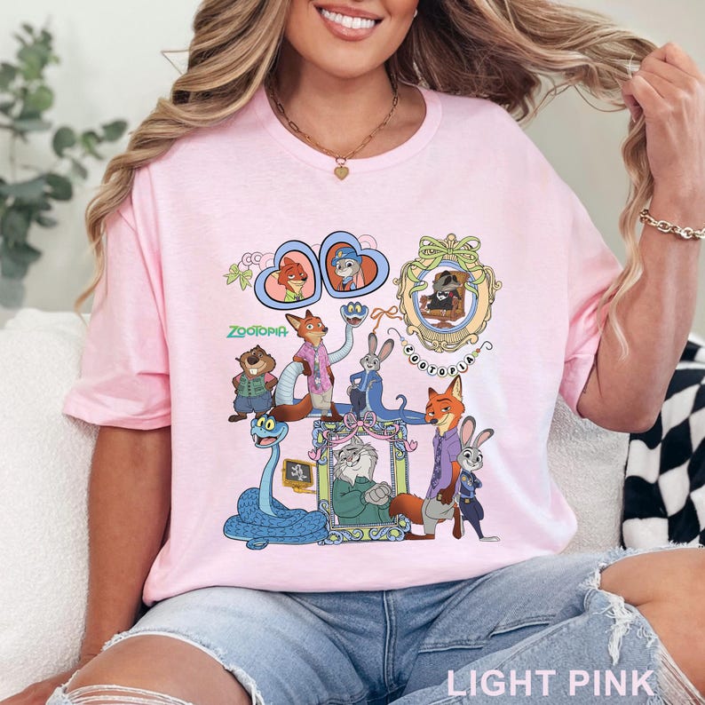 May include: Light pink t-shirt featuring a colorful graphic of characters from the animated film Zootopia. The design includes various animal characters in ornate frames and the word "Zootopia". The shirt is made of a soft material.