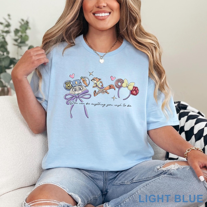 May include: A light blue t-shirt featuring a graphic print of a rabbit character, a fox character, and two sets of mouse ears. The text "You can be anything you wish to be" is printed in black script below the characters, accompanied by small hearts and stars.