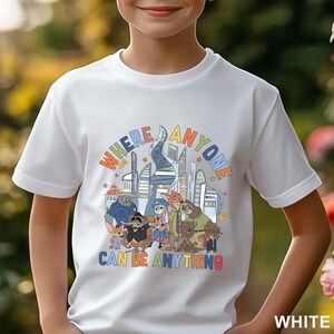 May include: White t-shirt featuring a colorful graphic with the text "WHERE ANYONE CAN BE ANYTHING" and cartoon characters. The design includes a cityscape and various animal characters. The shirt is short-sleeved.