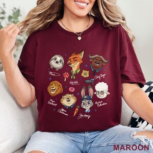 Puede incluir: Una camiseta de manga corta color granate con un estampado gr&aacute;fico de los personajes de Zootopia, incluyendo a Nick Wilde, Judy Hopps, Flash, Chief Bogo, Mayor Lionheart, Officer Clawhauser y Assistant Bellwether, junto a iconos y nombres.
