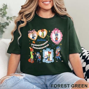 Pode incluir: Camiseta verde floresta com imagens de desenhos animados dos personagens de Zootopia. O design inclui imagens emolduradas e formas de cora&ccedil;&atilde;o, com "Zootopia" no centro. A camiseta &eacute; feita de um material macio. O texto "Forest Green" est&aacute; na parte inferior.