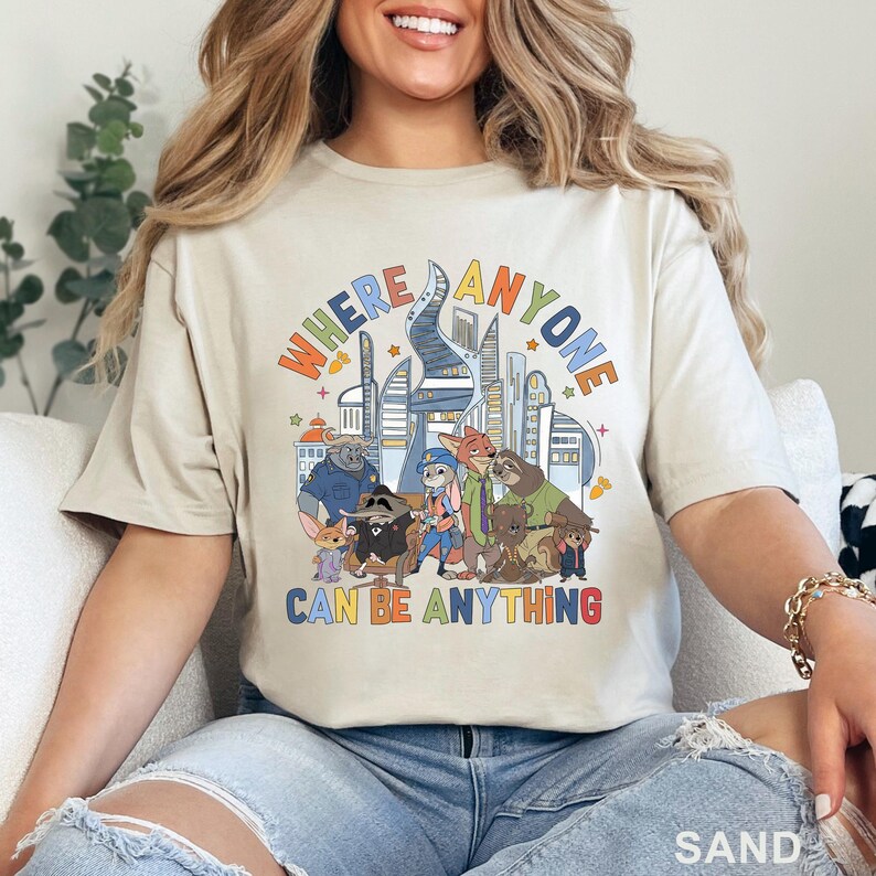 May include: Sand-colored t-shirt featuring a colorful graphic with the text "WHERE ANYONE CAN BE ANYTHING" above a cartoon cityscape and characters. The design includes various animal characters in a vibrant, playful style.