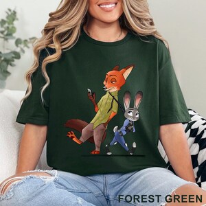 May include: Forest green t-shirt featuring a cartoon illustration of Nick Wilde and Judy Hopps from Zootopia. Nick, a fox, is holding a phone, while Judy, a rabbit, walks beside him. The text "FOREST GREEN" is at the bottom.