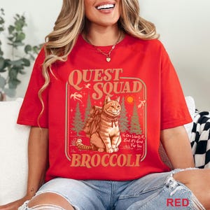 May include: Red t-shirt with a graphic featuring a cat wearing a backpack, the words "QUEST SQUAD" and "BROCCOLI". The design includes trees, a sun, and the text "No One Wants It, But It's Good For You".