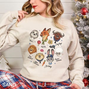 May include: A beige sweatshirt featuring cartoon characters from the movie Zootopia. The characters include Flash, Nick Wilde, Chief Bogo, Mayor Lionheart, Officer Clawhauser, Judy Hopps, and Assistant Bellwether. The word "SAND" is printed at the bottom.