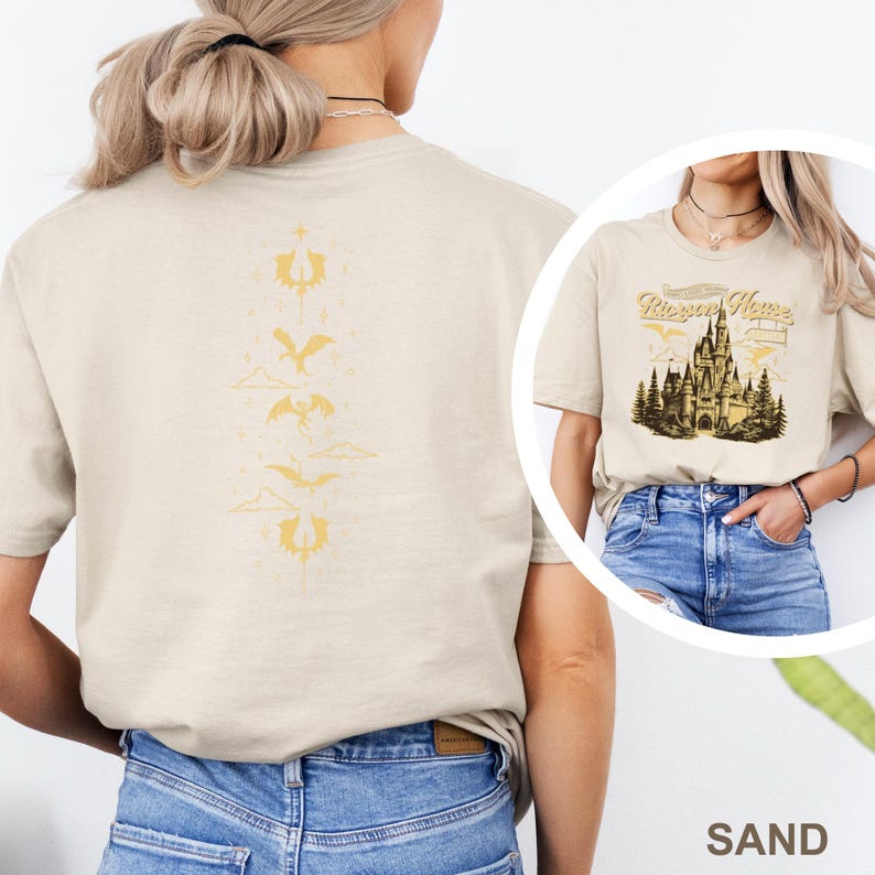 May include: Sand-coloured t-shirt featuring a castle graphic and the text "Rivera Home". The back of the shirt has a vertical design of gold-coloured stylized dragons and clouds. The t-shirt is made of a soft material.