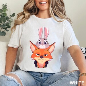 May include: White t-shirt with a cartoon illustration of a fox and a rabbit from the film Zootropolis. The fox is orange with a black collar, and the rabbit is grey with pink ears. The word "WHITE" is printed at the bottom.