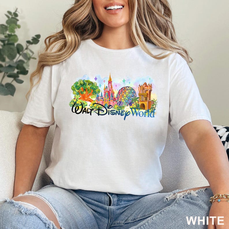May include: A white short-sleeved t-shirt featuring a colorful watercolor-style graphic of Walt Disney World landmarks, including a tree, a castle, a geodesic dome, and a tower, with the text Walt Disney World printed in a dark blue script font.