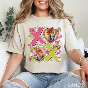 Peut inclure: Un t-shirt couleur sable avec une impression graphique de Nick Wilde et Judy Hopps de Zootopie. Le motif comprend de grandes lettres X et O roses et vertes, des cadres en forme de c&oelig;ur rose, des petits cadeaux, des cupcakes et des n&oelig;uds.