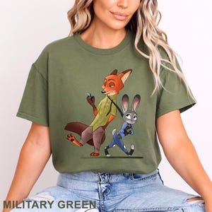 May include: Olive green t-shirt featuring a cartoon illustration of Nick Wilde and Judy Hopps from Zootopia. Nick, an orange fox, is holding a phone, while Judy, a gray rabbit, is walking. The text "MILITARY GREEN" is at the bottom.