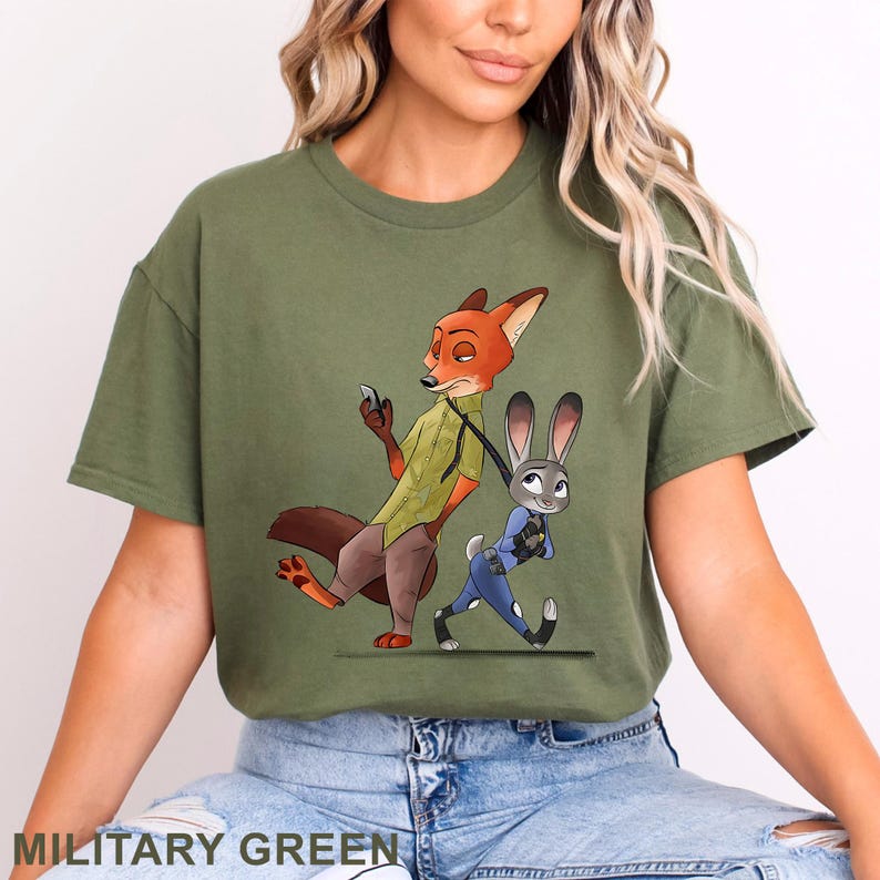 May include: Olive green t-shirt featuring a cartoon illustration of Nick Wilde and Judy Hopps from Zootopia. Nick, an orange fox, is holding a phone, while Judy, a gray rabbit, is walking. The text "MILITARY GREEN" is at the bottom.