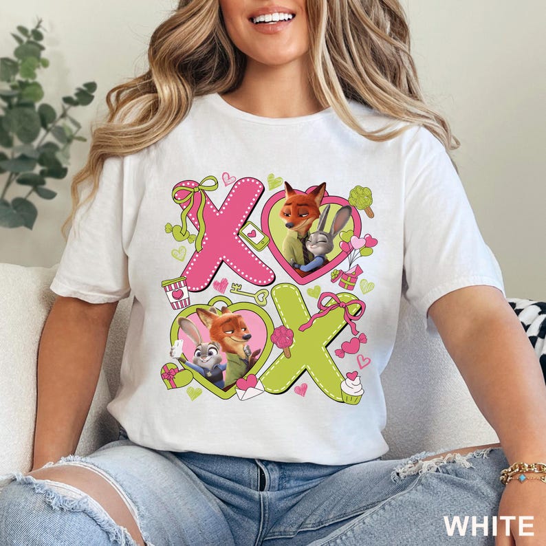 Peut inclure: Un t-shirt blanc avec une impression graphique affichant le texte XOXO. Le motif comprend des lettres roses et vert citron, des c&oelig;urs et des illustrations de Nick Wilde et Judy Hopps de Zootopie, accompagn&eacute;s de petits c&oelig;urs et cadeaux.