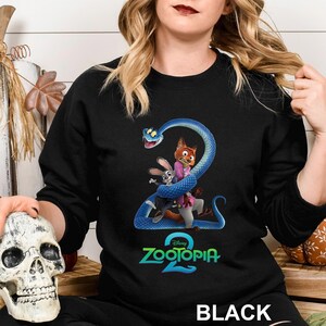 May include: Black crewneck sweatshirt featuring a colorful graphic from the Disney movie Zootopia 2. The design includes the characters Nick Wilde, Judy Hopps, and a blue snake. The word "BLACK" is printed at the bottom.
