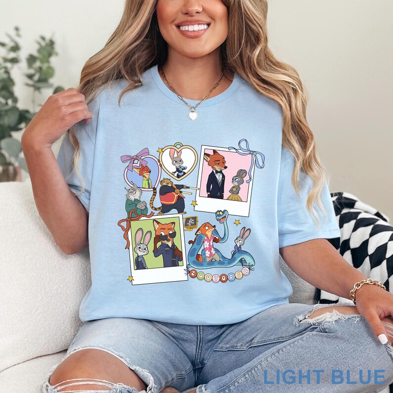 May include: A light blue t-shirt featuring a graphic print of Zootopia characters including Judy Hopps and Nick Wilde. The design includes multiple framed illustrations, polaroid-style photos, and the word Zootopia in a colorful bubble font.