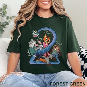 Puede incluir: Una camiseta de color verde bosque oscuro con un estampado gr&aacute;fico de personajes de dibujos animados, incluyendo un zorro, un conejo, un lince, un castor y una serpiente. El texto "Judy", "Nick", "Nibbles" y "Pawbert" aparece en blanco.