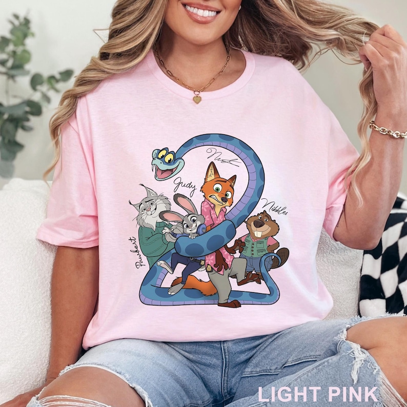 Puede incluir: Una camiseta rosa claro con un estampado gr&aacute;fico de una serpiente azul enroscada alrededor de un grupo de personajes de dibujos animados, incluyendo un zorro, un conejo, un lince y un castor. Los nombres Pawbert, Judy, Nick y Nibbles est&aacute;n escritos.