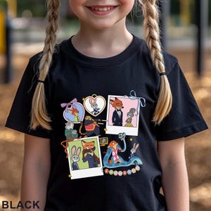 May include: A black short-sleeved t-shirt featuring a graphic print of Zootopia characters. The design includes multiple framed illustrations of Nick Wilde and Judy Hopps, along with other characters, arranged in a collage style with the text Zootopia.