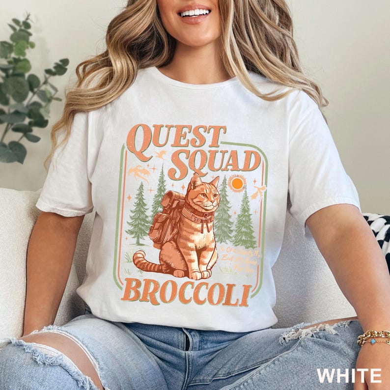 May include: White t-shirt with a graphic featuring a cat wearing a backpack, the words "QUEST SQUAD BROCCOLI", trees, and a sun. The design is in orange and green. The shirt is labeled "WHITE" at the bottom.