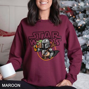 May include: Maroon sweatshirt featuring a Star Wars graphic. The design includes the Star Wars logo above an illustration of The Mandalorian and Grogu. The Mandalorian is in silver armor, holding Grogu. The sweatshirt is a solid maroon color.