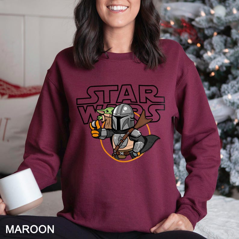 May include: Maroon sweatshirt featuring a Star Wars graphic. The design includes the Star Wars logo above an illustration of The Mandalorian and Grogu. The Mandalorian is in silver armor, holding Grogu. The sweatshirt is a solid maroon color.