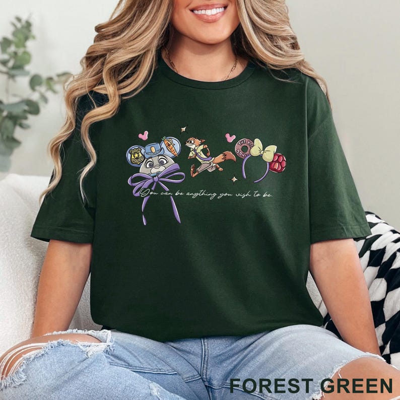 May include: A forest green short-sleeve t-shirt featuring a graphic of a rabbit character with a police badge, a fox character, and two sets of mouse ears. The text "You can be anything you wish to be" is printed in white script below the characters.