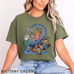 Puede incluir: Una camiseta verde oliva con un estampado gr&aacute;fico de personajes de dibujos animados, incluyendo un zorro, un conejo, un lince, un castor y una serpiente azul. Los nombres Judy, Nick, Pawbert y Nibbles est&aacute;n escritos en cursiva.