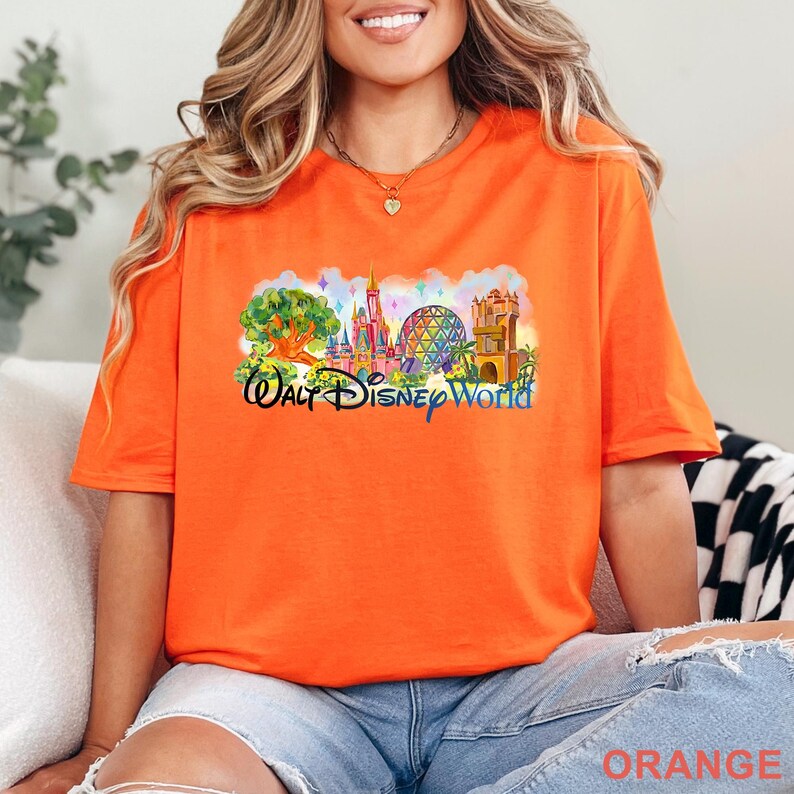 May include: A bright orange short-sleeve t-shirt featuring a colorful graphic print of the Walt Disney World skyline, including a castle, a geodesic dome, and a tree. The text Walt Disney World is printed in a dark blue script font below the icons.