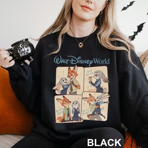 Puede incluir: Sudadera negra con un dise&ntilde;o de dibujos animados de los personajes de Zootopia. El dise&ntilde;o incluye cuatro paneles con Nick Wilde y Judy Hopps en varias poses, con las palabras "Walt Disney World" encima de los paneles y "BLACK" debajo.