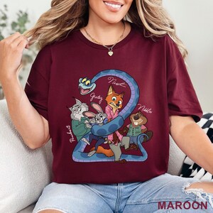 Puede incluir: Una camiseta color granate con un estampado gr&aacute;fico de personajes de dibujos animados, incluyendo un zorro, un conejo, una serpiente, un lince y un castor. Los nombres Pawbert, Judy, Nick y Nibbles est&aacute;n escritos en letra blanca.