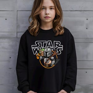 May include: Black sweatshirt featuring a Star Wars graphic. The design includes the text "STAR WARS" in white, along with an illustration of The Mandalorian and Grogu. The Mandalorian is depicted in silver armor, holding a weapon.