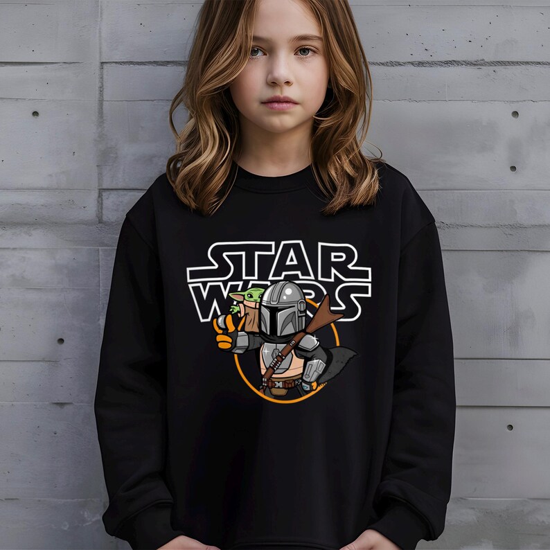 May include: Black sweatshirt featuring a Star Wars graphic. The design includes the text "STAR WARS" in white, along with an illustration of The Mandalorian and Grogu. The Mandalorian is depicted in silver armor, holding a weapon.