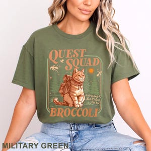 May include: Military green t-shirt with the text "QUEST SQUAD BROCCOLI". The graphic features a cartoon cat with a backpack in a forest setting, with the text "No One Wants It, But It's Good For You".