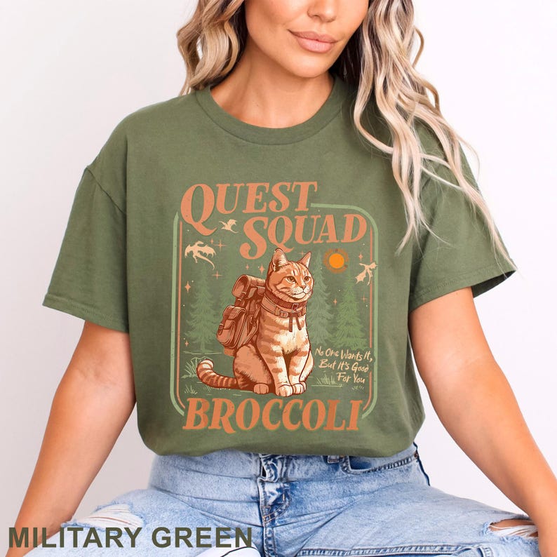 May include: Military green t-shirt with the text "QUEST SQUAD BROCCOLI". The graphic features a cartoon cat with a backpack in a forest setting, with the text "No One Wants It, But It's Good For You".