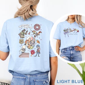 May include: A light blue t-shirt featuring a graphic print of animated characters from Zootopia, including Nick Wilde and Judy Hopps, alongside icons like a donut, a badge, and a carrot. The text reads, "You can be whatever you wish to be."