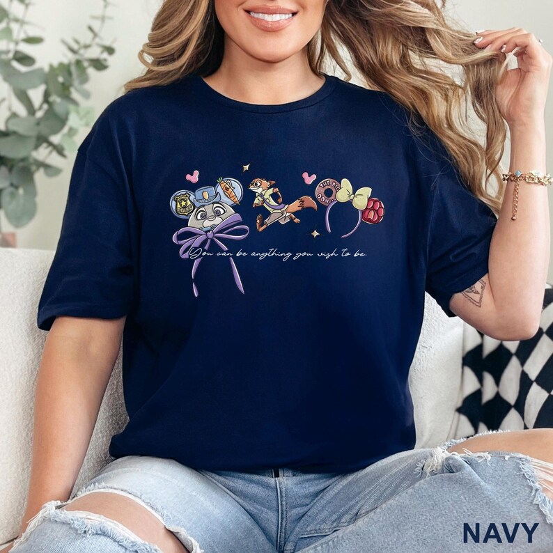 May include: A navy blue t-shirt featuring a graphic print of a rabbit character wearing a police hat, a fox character, and two sets of mouse ears. The text "You can be anything you wish to be" is printed in white script below the colorful illustrations.