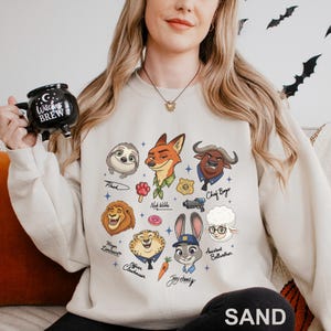May include: Sand-coloured sweatshirt with cartoon illustrations of Zootopia characters, such as Nick Wilde, Judy Hopps, and Flash. The sweatshirt has a crew neck and long sleeves. The word "SAND" is printed at the bottom.