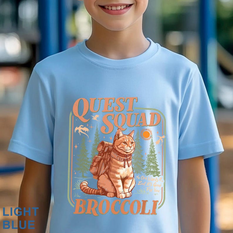 May include: Light blue t-shirt with a graphic featuring an orange tabby cat wearing a backpack, the words "QUEST SQUAD" and "BROCCOLI". The design includes trees, a sun, and a dragon.