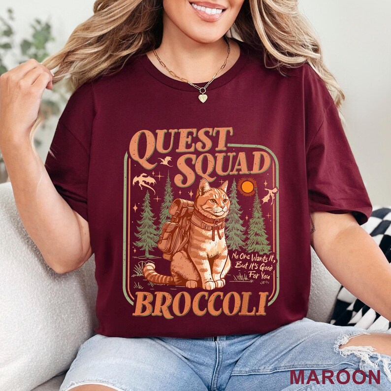 May include: Maroon t-shirt with a graphic featuring a cat with a backpack, the words "Quest Squad" and "Broccoli". The design includes trees, a sun, and the text "No One Wants It, But It's Good For You."