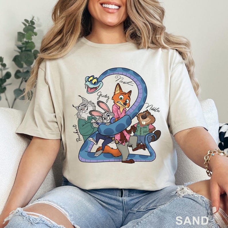 Puede incluir: Una camiseta color arena con un estampado gr&aacute;fico de cinco personajes de dibujos animados: una serpiente azul, un gato, un conejo, un zorro y un castor. Los personajes est&aacute;n agrupados con nombres escritos a mano como Pawbert, Judy, Nick y Nibbles.