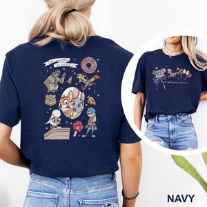 May include: A navy blue t-shirt featuring a Zootopia-themed graphic print. The back displays various characters, a badge, a donut, and a carrot, with the text "You can be whatever you wish to be." The front shows smaller character icons and ears.