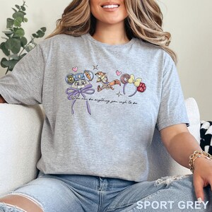 May include: A heather gray short-sleeve t-shirt featuring a graphic print of two cartoon characters, mouse ears, a donut, and the text "You can be anything you wish to be." The design includes small pink hearts and stars in a colorful illustration.