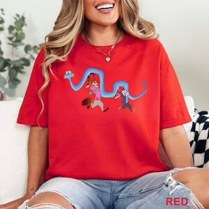 May include: A bright red short-sleeved t-shirt featuring a graphic print of a long blue snake held up by a cartoon fox wearing a pink shirt and a cartoon rabbit wearing a blue shirt. The design is centered on the chest of the crew neck garment.