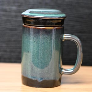 May include: A ceramic mug with a lid, featuring a teal and brown glaze. The mug has a handle and a cylindrical shape. The lid is flat and fits snugly on top. The mug is approximately 5 inches tall.