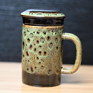 May include: A ceramic mug with a lid, featuring a speckled green and brown glaze. The mug has a handle and a dark brown base and lid. The mug is approximately 5 inches tall.