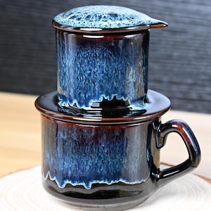 May include: A ceramic coffee filter set with a dark blue and brown glaze. The set includes a mug with a handle, a base, and a filter. The glaze has a dripped effect, and the set is on a wooden coaster.