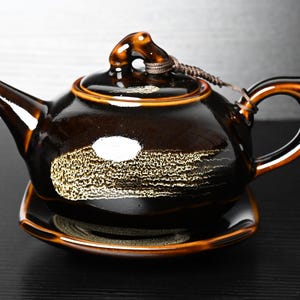 Puede incluir: Una tetera de cerámica esmaltada en color marrón oscuro con un plato cuadrado a juego. La tetera presenta un acabado brillante, un pico curvo, un asa y una tapa asegurada con un cordón marrón. Un diseño dorado texturizado adorna el lateral.