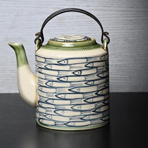 May include: A cylindrical ceramic teapot featuring a repeating blue hand-painted fish pattern on a cream-coloured body. The teapot has a light green glazed spout, a matching lid, and a dark metal swing handle attached to two small metal loops.