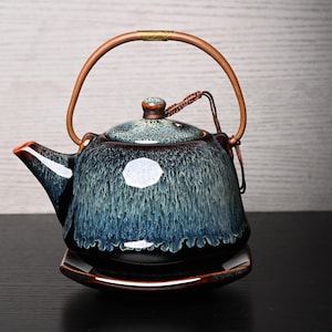 May include: A ceramic teapot with a glossy glaze featuring a gradient of deep black at the base transitioning into a mottled blue and white finish. It includes a curved handle, a matching lid with a small knob, and a square-shaped base plate.